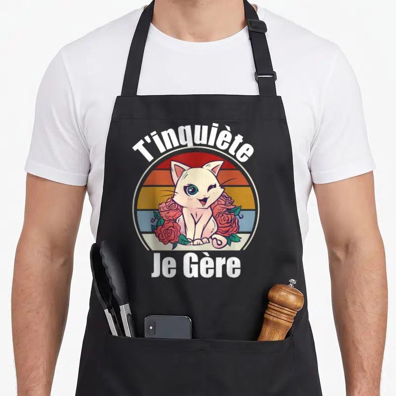 Tablier de cuisine noir avec un chat blanc kawaii entouré de roses et le texte T'inquiète Je Gère en style rétro.
