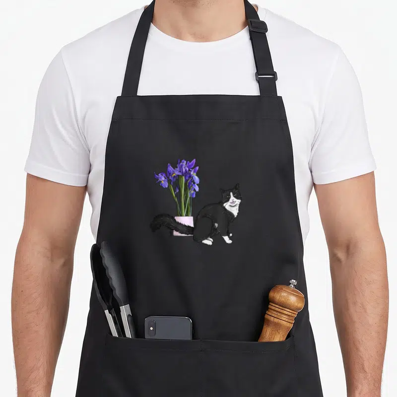 Tablier de cuisine noir avec un chat noir et blanc souriant près d'un pot rose aux iris violets