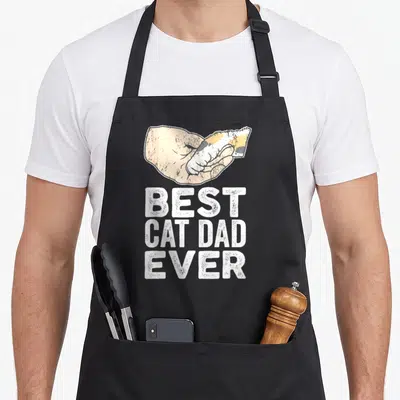 Tablier de cuisine noir avec une main tenant un chaton et le texte BEST CAT DAD EVER en lettres blanches.