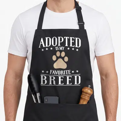 Tablier de cuisine noir avec inscription Adopted Is My Favorite Breed et patte d'animal beige au centre.