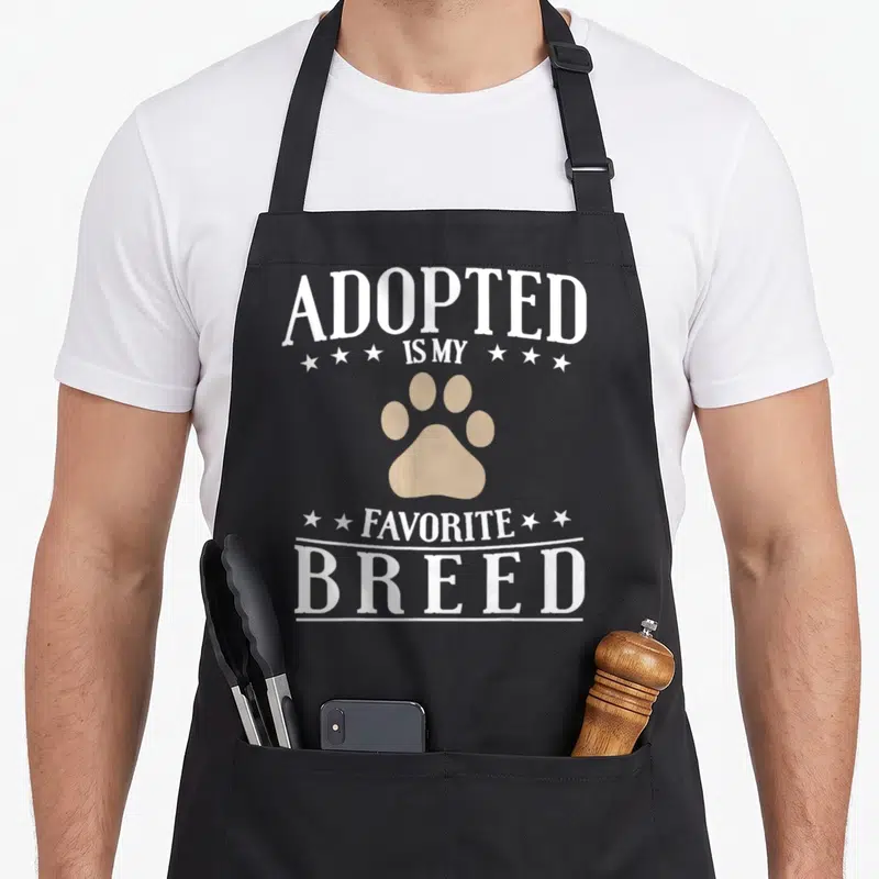 Tablier de cuisine noir avec inscription Adopted Is My Favorite Breed et patte d'animal beige au centre.