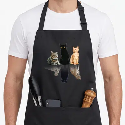 Tablier de cuisine noir avec trois chats aux reflets de guépard et tigre imprimés sur le devant.