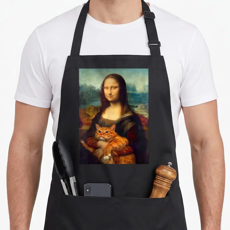 Tablier de cuisine noir avec une illustration de la Mona Lisa tenant un chat roux dans ses bras sur fond de paysage flou.