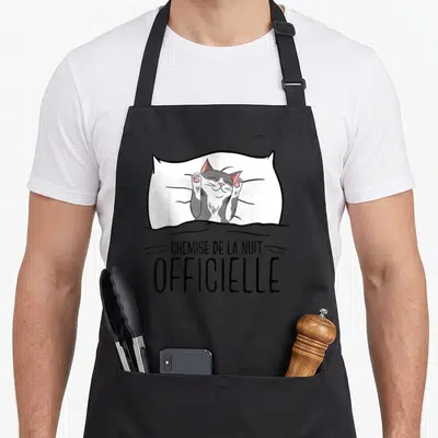 Tablier de cuisine noir avec illustration d'un chat gris et blanc s'étirant dans un oreiller blanc au-dessus du texte « chemise de la nuit ».