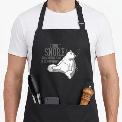 Tablier de cuisine noir avec un chat blanc allongé et le texte I don't snore I purr with the force of 10,000 kittens en gris.
