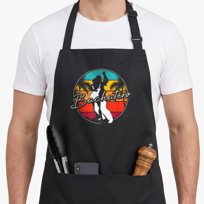 Tablier de cuisine noir avec silhouette de danseur Bachatero et texte cursive sur fond de soleil tropical.