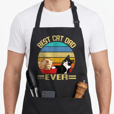 Tablier de cuisine noir avec illustration vintage Best Cat Dad Ever montrant un poing et un chat bicolore.
