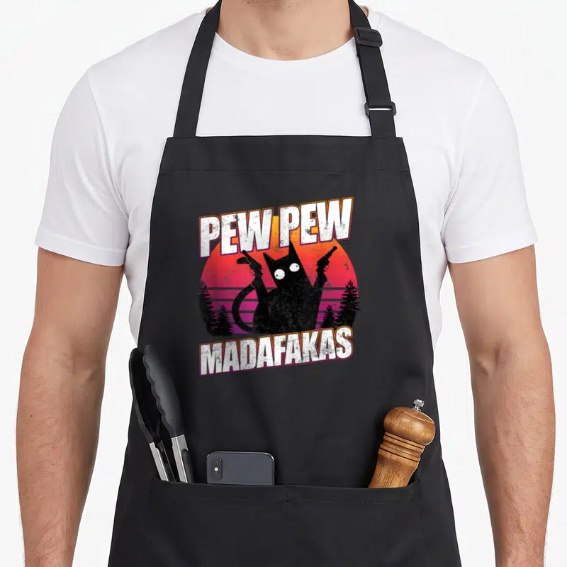 Tablier de cuisine noir avec illustration d'un chat noir tenant deux revolvers sur fond dégradé rose et orange.