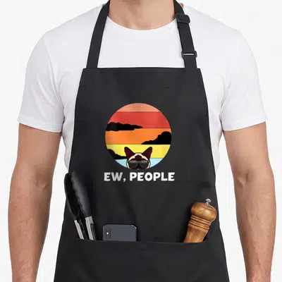 Tablier de cuisine noir avec illustration d'un chat Siamese en lunettes noires devant un soleil couchant coloré et le texte Ew, Humains.