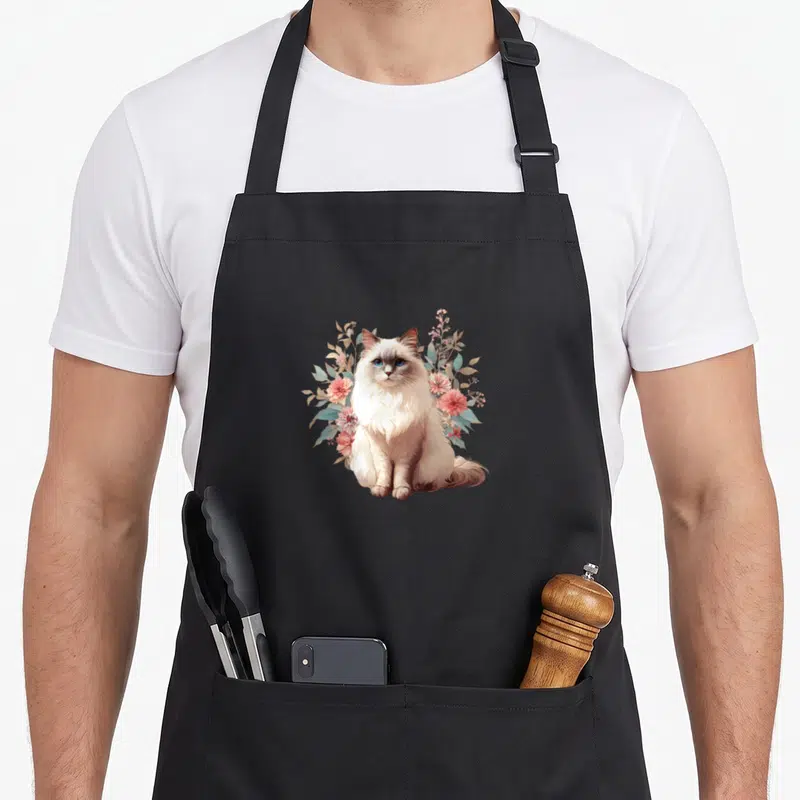 Tablier de cuisine noir avec illustration d'un chat Ragdoll aux yeux bleus entouré de fleurs roses et de feuilles vertes.