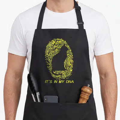 Tablier de cuisine noir avec une empreinte digitale jaune stylisée en forme de chat et le texte It's In My DNA en bas.