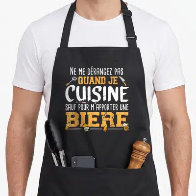Tablier de cuisine noir avec texte Ne me dérangez pas quand je cuisine sauf pour m'apporter une bière entouré d'ustensiles et de bouteilles.