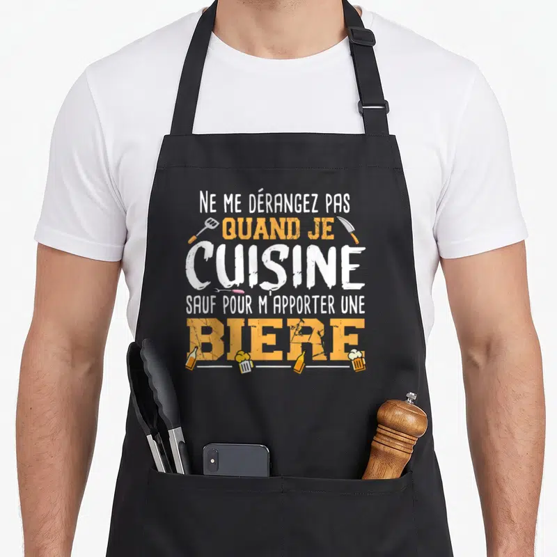 Tablier de cuisine noir avec texte Ne me dérangez pas quand je cuisine sauf pour m'apporter une bière entouré d'ustensiles et de bouteilles.
