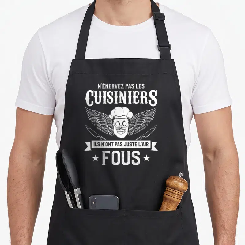 Tablier de cuisine noir avec un crâne de cuisinier aux ailes, couteaux croisés et texte humoristique en blanc.