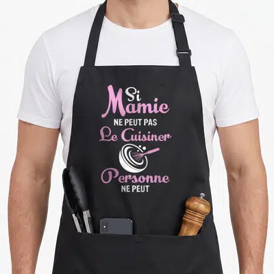 Tablier de cuisine noir avec texte rose et blanc Si Mamie Ne Peut Pas Le Cuisiner Personne Ne Peut et un fouet dans un bol.