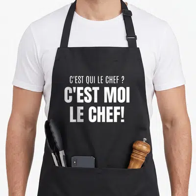 Tablier de cuisine noir avec inscription blanche C'EST QUI LE CHEF C'EST MOI LE CHEF en typographie audacieuse et contrastée.