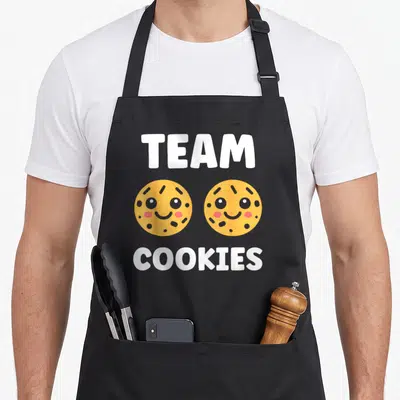 Tablier de cuisine noir avec l'inscription Team Cookies et deux biscuits jaunes aux visages ronds et joyeux au centre.