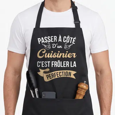 Tablier de cuisine noir avec texte blanc et doré et deux poêles stylisées.