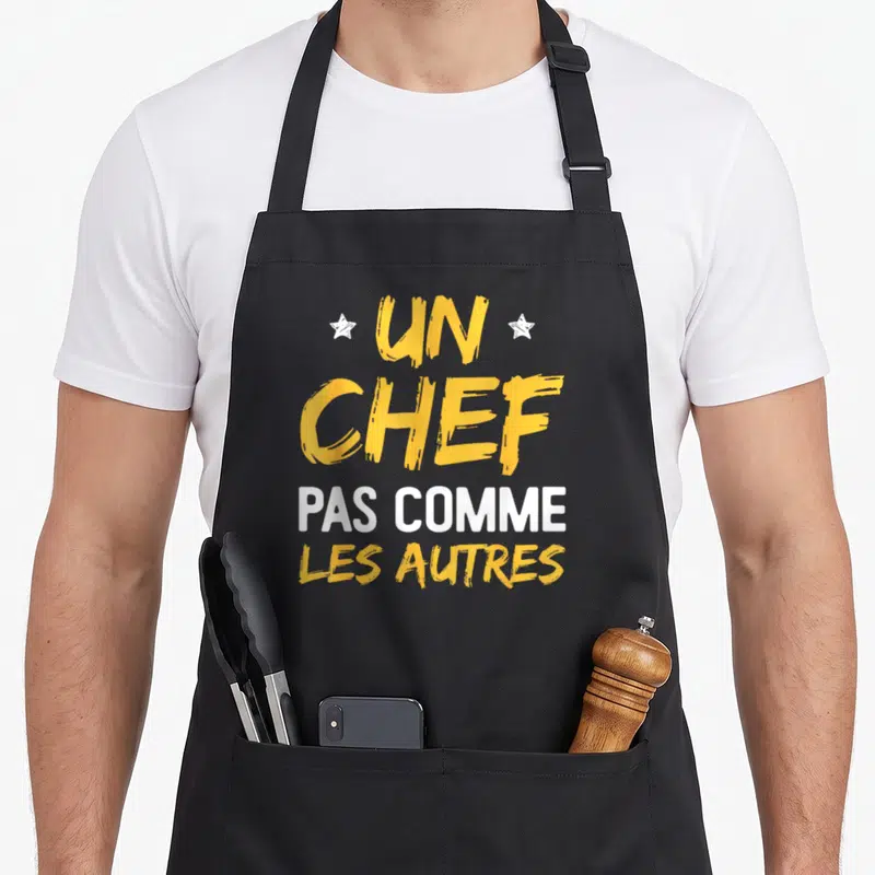 Tablier de cuisine noir avec inscription Un Chef Pas Comme Les Autres en lettres jaunes et blanches ornées d'étoiles.