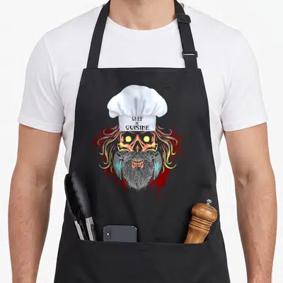 Tablier de cuisine noir avec un crâne de cuisinier à la toque blanche, barbe noire et flammes colorées.