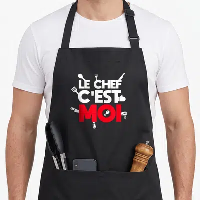Tablier de cuisine noir avec l'inscription Le Chef C'est Moi en blanc et rouge, orné d'icônes d'ustensiles culinaires blancs.