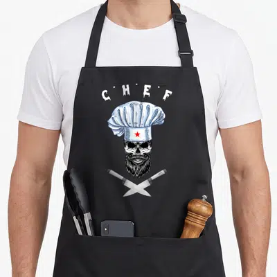 Tablier de cuisine noir avec crâne barbu, toque blanche et couteaux croisés