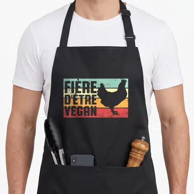 Tablier de cuisine noir avec motif vintage affichant une silhouette de poulet sur fond de bandes colorées et le texte fière d'être vegan.