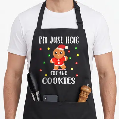 Tablier de cuisine noir avec un biscuit de Noël en chapeau rouge et texte blanc humoristique.