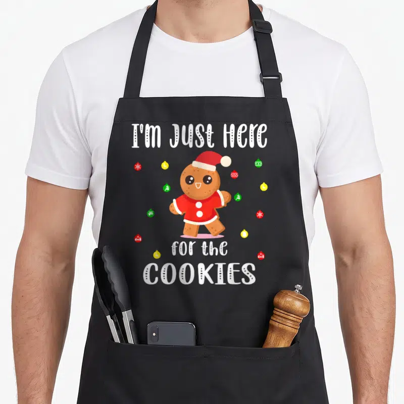 Tablier de cuisine noir avec un biscuit de Noël en chapeau rouge et texte blanc humoristique.