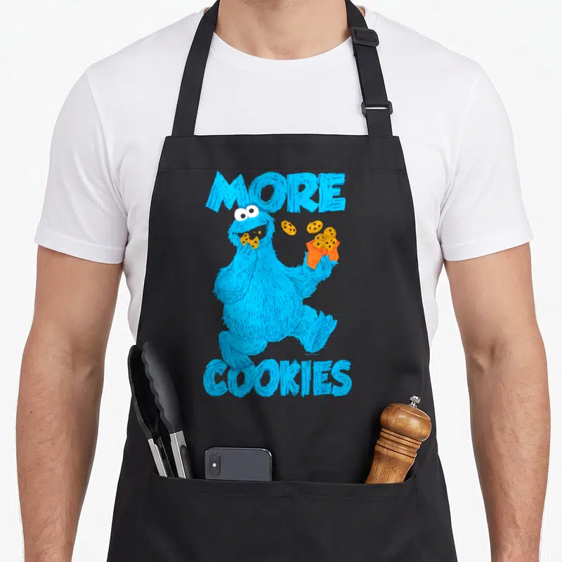 Tablier de cuisine noir avec un monstre bleu vorace tenant des biscuits et le texte plus de cookies en lettres capitales bleues.