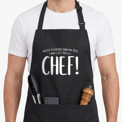 Tablier de cuisine noir avec inscription blanche humoristique affirmant que l'on est plusieurs dans la tête mais que l'on est le chef.