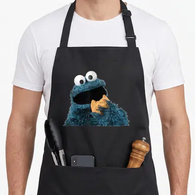 Tablier de cuisine noir avec Cookie Monster bleu tenant un biscuit croqué dans sa main fourrue.