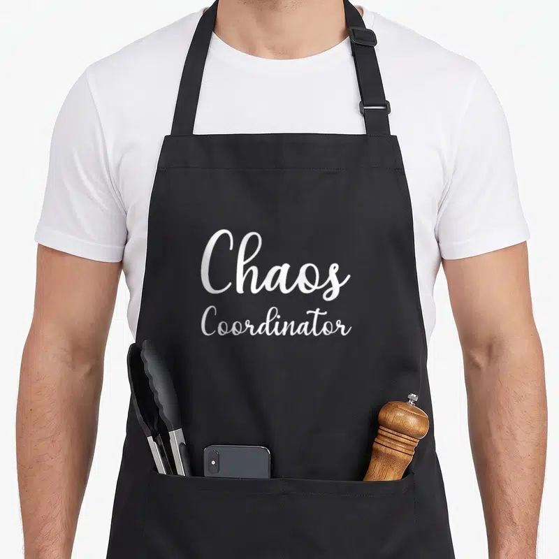 Tablier de cuisine noir avec l'inscription blanche Chaos Coordinator en écriture cursive élégante au centre.