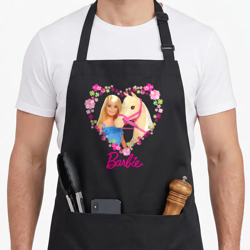Tablier de cuisine noir avec Barbie et un cheval blond dans un cœur de fleurs roses et le logo Barbie en bas.