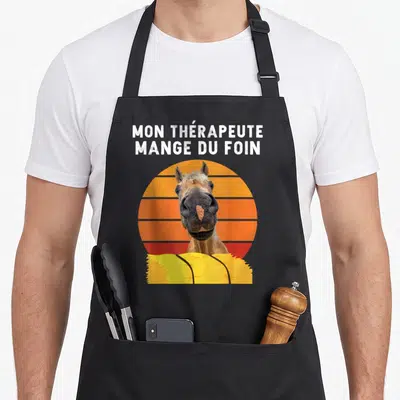 Tablier de cuisine noir avec inscription mon thérapeute mange du foin et portrait de cheval sur fond orange rayé.