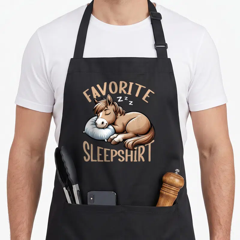 Tablier de cuisine noir avec illustration d'un poney marron dormant sur un coussin blanc sous le texte Favorite Sleepshirt en beige.