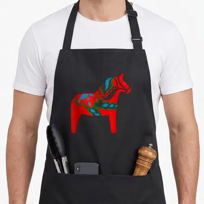 Tablier de cuisine noir avec un grand cheval Dala rouge aux décors floraux bleus et verts typiques de l'artisanat suédois.