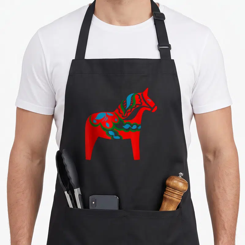 Tablier de cuisine noir avec un grand cheval Dala rouge aux décors floraux bleus et verts typiques de l'artisanat suédois.