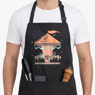 Tablier de cuisine noir avec illustration colorée d'un carrousel vintage et de chevaux en mouvement sous une bannière étoilée.