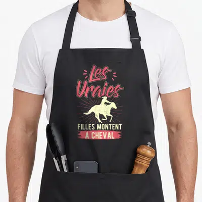 Tablier de cuisine noir avec illustration d'une cavalière sur un cheval au galop et texte Les Vraies Filles Montent À Cheval en rose et crème.