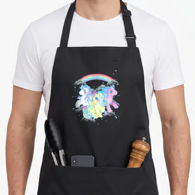 Tablier de cuisine noir avec trois poneys colorés sous un arc en ciel Magie et étoiles scintillantes autour du motif central.