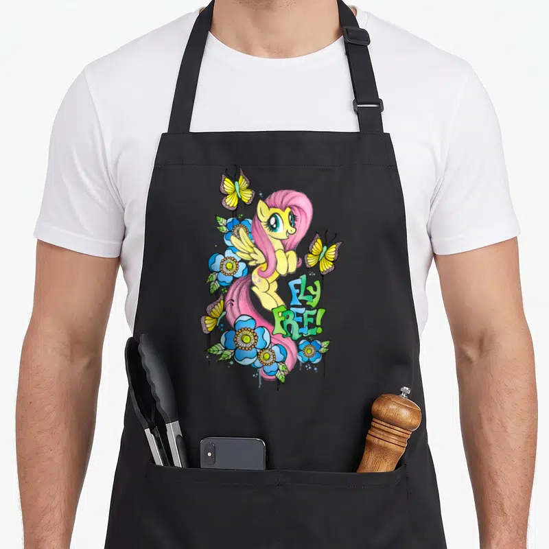 Tablier de cuisine noir avec Fluttershy, des papillons jaunes et des fleurs bleues autour du texte Fly Free en lettres stylisées.