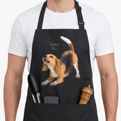 Tablier de cuisine noir avec un beagle en posture de jeu et le texte Love is... imprimé en rose.
