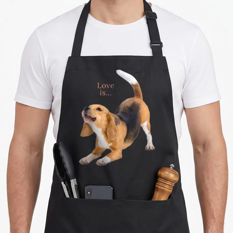 Tablier de cuisine noir avec un beagle en posture de jeu et le texte Love is... imprimé en rose.
