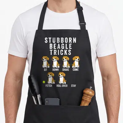 Tablier de cuisine noir avec six beagles illustrés sous les mots Stubborn Beagle Tricks et leurs commandes anglaises.