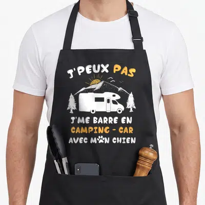Tablier de cuisine noir avec inscription J'peux pas j'me barre en camping-car avec mon chien et un camping-car blanc sur fond de montagne.