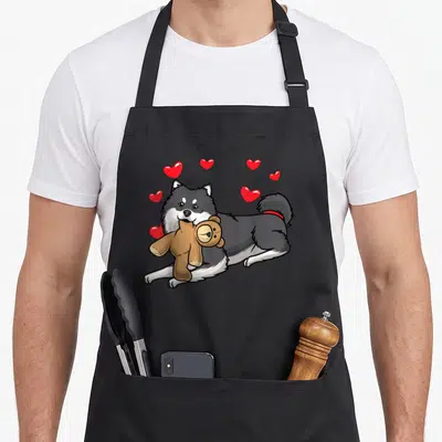 Tablier de cuisine noir avec un chien finnois de Laponie tenant une peluche et des cœurs rouges flottants.