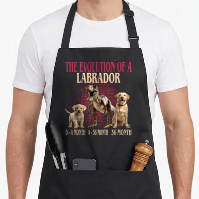 Tablier de cuisine noir avec une illustration montrant un chiot, un T-Rex et un labrador adulte sur fond rose.