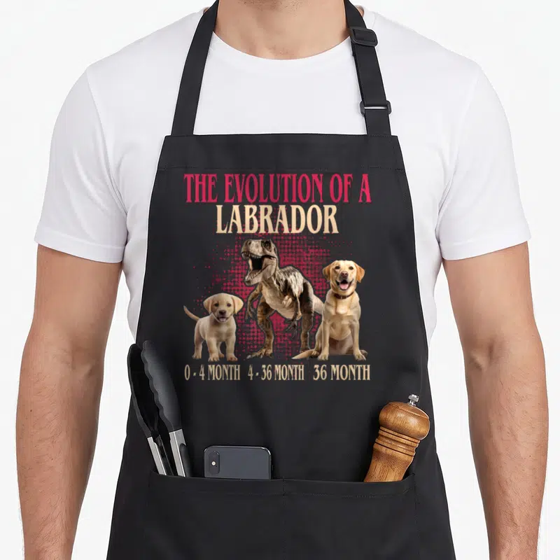 Tablier de cuisine noir avec une illustration montrant un chiot, un T-Rex et un labrador adulte sur fond rose.