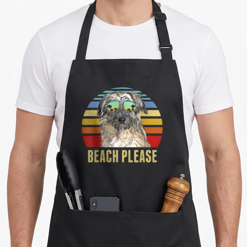 Tablier de cuisine noir avec illustration d'un chien en lunettes de soleil et texte Beach Please sur fond de soleil coloré.